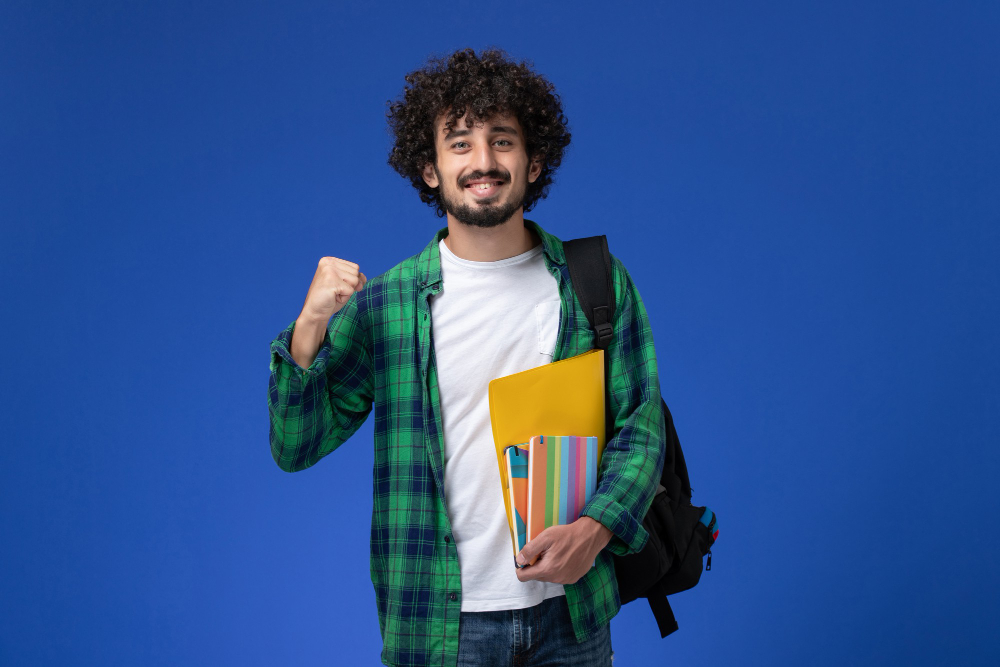 vista frontal de um estudante do sexo masculino usando uma mochila preta segurando cadernos e arquivos na parede azul
